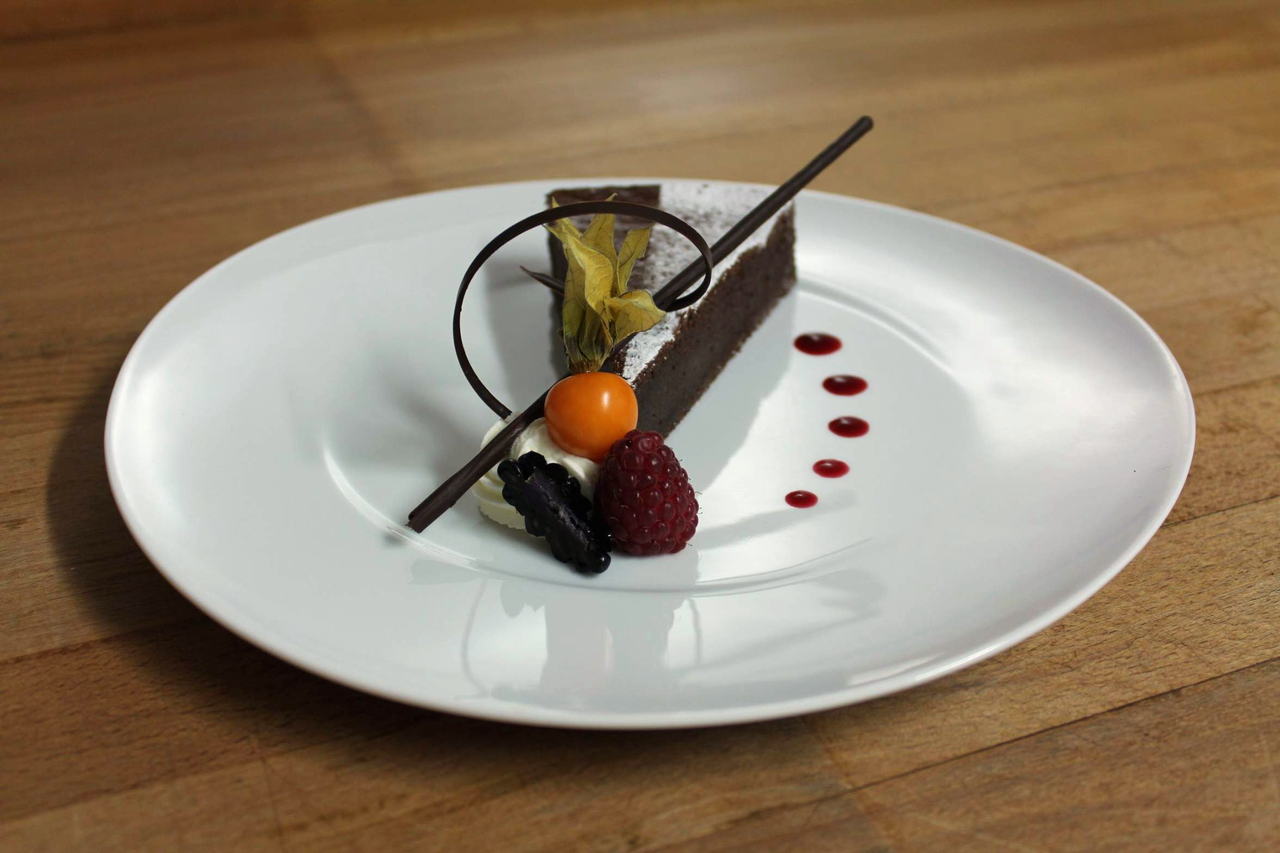 Tellerdessert: Schokoladenschnitte mit Physalis, Himbeere und Brombeere, dazu Schokoladendekor und Fruchtsauce auf weissem Teller